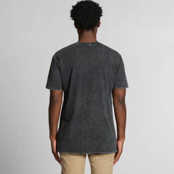 A male model with dark skin and curly hair is shown from behind, wearing a black stone-washed T-shirt with visible stitching details and beige trousers. The T-shirt has a relaxed fit and a slightly faded look.