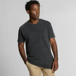 A male model with dark skin and curly hair is standing facing the camera, wearing a black stone-washed T-shirt and beige trousers. The T-shirt has a stone wash effect and a casual, classic crew neck fit.
