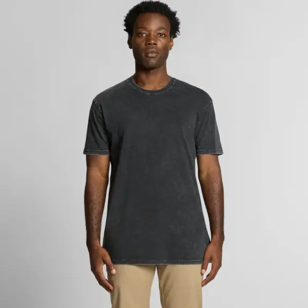 A male model with dark skin and curly hair is standing facing the camera, wearing a black stone-washed T-shirt and beige trousers. The T-shirt has a slightly faded effect and a casual, classic crew neck fit.