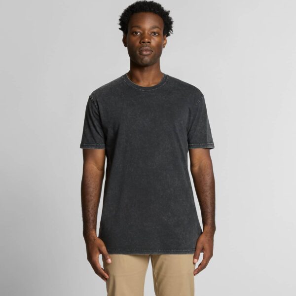 A male model with dark skin and curly hair is standing facing the camera, wearing a black stone-washed T-shirt and beige trousers. The T-shirt has a slightly faded effect and a casual, classic crew neck fit.
