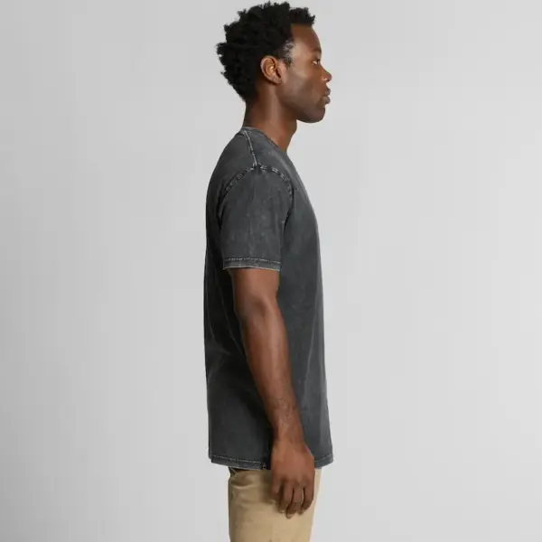 A male model with dark skin and curly hair is standing sideways, wearing a black stone-washed T-shirt and beige trousers. The T-shirt has a slightly faded effect and a casual, classic crew neck fit.