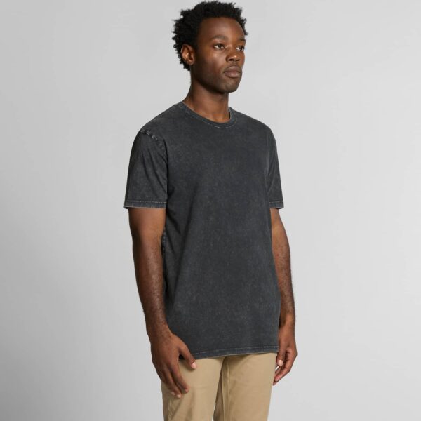 A male model with dark skin and curly hair wears a vintage-inspired black T-shirt with a faded stone-wash finish. Styled with beige trousers, perfect for casual and streetwear outfits.