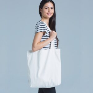 person carrying a plain white tote bag on their right shoulder, standing sideways to the camera in front of a plain blue background