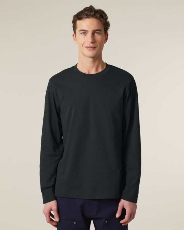 man in black long sleeve stanley stella creator shirt and black trousers in front of a plain beige background.