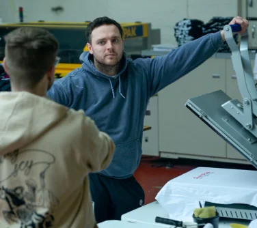 a man standing by a dtf heat press machine in a clothing printing warehouse.