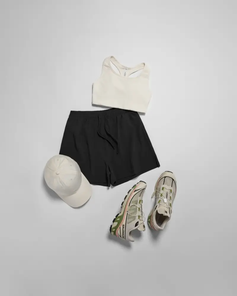 flat lay of activewear by brand 'as colour', showing a cream-coloured sports bra, black shorts, a cream-coloured cap and branded trainers in front of a light grey background.