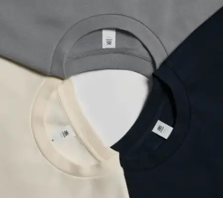 Grey, ecru and black AS Colour T-shirts in a circle, connected by their collars to showcase detail.
