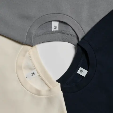 Grey, ecru and black AS Colour T-shirts in a circle, connected by their collars to showcase detail.