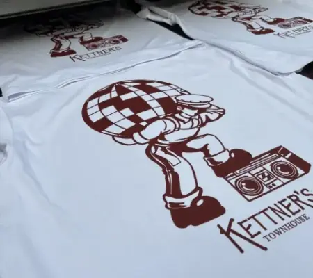 Multiple white t-shirts with a screen printed disco style burgundy design, coming off of an industrial tunnel dryer.
