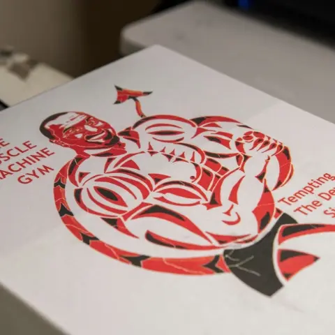 A digital printing design on a white t shirt of a devil with text that reads "muscle machine gym. tempting the devil since 1890"