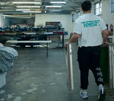 Teesh team member carrying screen printing frames across a professional print studio, with stacks of garments and automated machinery in the background.