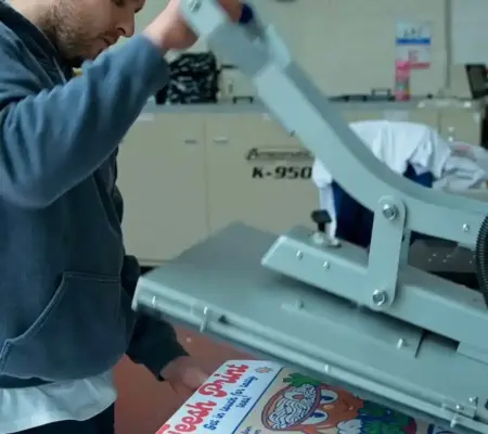 A garment printing professional using a heat press to apply a colorful transfer design onto a white T-shirt in a workshop setting.